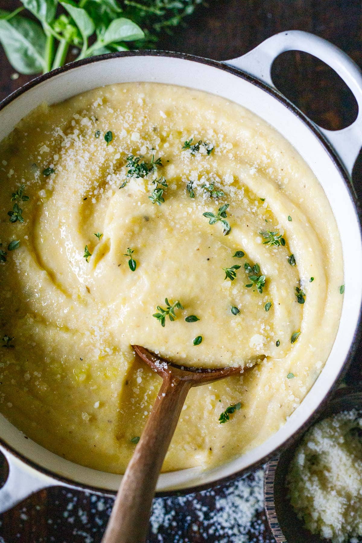 large pot of creamy polenta topped with parmesan and fresh herbs with wooden spoon.